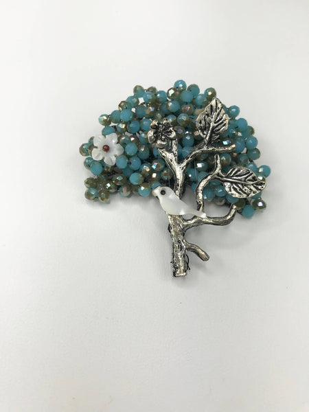 Silver-tone tree brooch with intricate branches, a white bird, floral details, and sparkling teal crystal beads. Lightweight and durable, available in white and green for versatile styling.